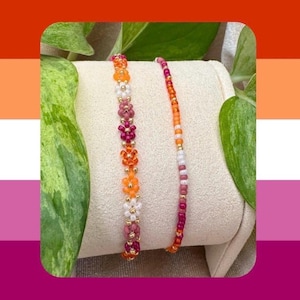 May include: Two beaded bracelets displayed on a curved stand. One bracelet features a repeating pattern of small orange, pink, and white flower-shaped beads with gold accents. The second bracelet consists of a single strand of small round beads.