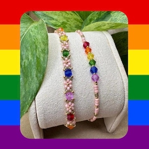 May include: Two beaded bracelets featuring light pink seed beads. One bracelet displays a repeating flower pattern with multi-colored crystal accents in yellow, green, blue, purple, and red. The second bracelet features a single row of rainbow crystals.