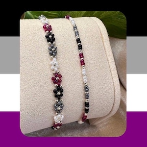 May include: Two beaded bracelets. One features flower-shaped beads in white, black, gray, and burgundy. The other bracelet has a linear design with black, burgundy, gray, and white beads. Both bracelets are displayed on a beige surface.