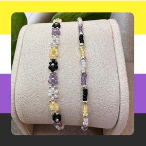May include: Two beaded bracelets featuring small, round glass beads. The left bracelet displays a repeating floral pattern in black, white, yellow, and purple. The right bracelet consists of a single strand of alternating black, white, yellow, and purple beads.