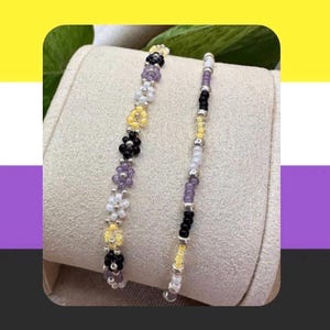 May include: Two beaded bracelets. One features flower-shaped beads in black, yellow, white, and purple. The other bracelet has a linear design with black, yellow, white, and purple beads. Both bracelets are on a neutral background.
