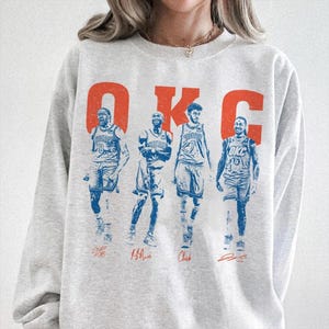 Op de afbeelding: Een lichtgrijs sweatshirt met een afbeelding van vier basketballers in blauwe en witte uniformen. Het woord "OKC" is in oranje boven de spelers gedrukt. De namen van de spelers staan eronder in een scriptletter.