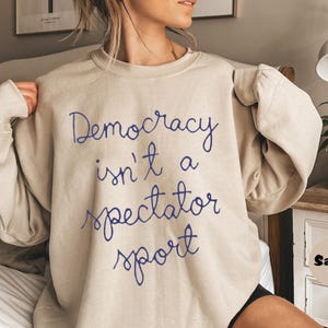 Può includere: Felpa beige con la scritta blu "Democracy isn't a spectator sport". Il top a maniche lunghe è realizzato in materiale morbido. La felpa è in stile casual.
