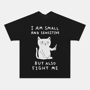 以下が含まれることがあります： 黒いTシャツに、ナイフを持った猫の白いイラスト。Tシャツのテキストは「I AM SMALL AND SENSITIVE BUT ALSO FIGHT ME」と書かれています。猫のイラストはミニマリストデザインです。