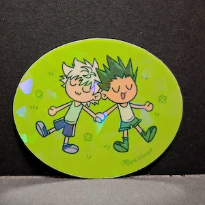 May include: A holographic sticker depicting two cartoon characters holding hands on a green field. One character has white hair and blue shorts, the other has green hair and a green outfit. The sticker has a shiny, iridescent finish.