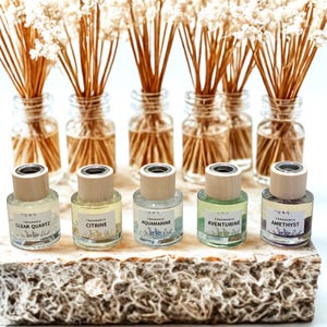 May include: A selection of small glass bottles with wooden lids, each labelled with a gemstone name, including "Clear Quartz" and "Citrine." The bottles are arranged on a textured surface, with small jars of dried flowers in the background.