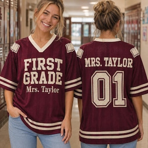 Puede incluir: Una camiseta de malla color granate con ribetes y rayas color crema en las mangas y el dobladillo. El frente presenta el texto "FIRST GRADE" y "Mrs. Taylor" con iconos de libros en los hombros. La espalda muestra "MRS. TAYLOR" y el número "01".