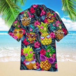 Op de afbeelding: Een overhemd met korte mouwen en knoopsluiting met een levendige tropische print van gele ananassen die verschillende zonnebrillen dragen, afgewisseld met felroze hibiscusbloemen en donkergroene palmbladeren op een zwarte achtergrond.
