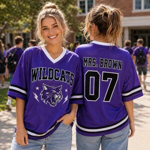 Puede incluir: Una camiseta de malla morada con cuello en V blanco y mangas cortas con rayas negras y blancas. El frente muestra el texto WILDCATS sobre un gráfico de una cabeza de gato montés. La parte posterior presenta el texto MRS. BROWN y el número 07.