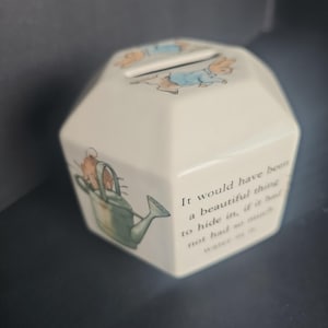 May include: A cream-colored ceramic money box with an octagonal shape. The top features a coin slot and a rabbit illustration. The side displays a green watering can with a rabbit and the text: It would have been a beautiful thing to hide in, if it had not had so much water in it.