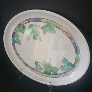 May include: An oval white serving tray featuring a decorative border with a geometric pattern in shades of purple, orange, and green. The center of the tray is decorated with green ivy leaves positioned at the top left and bottom right corners.