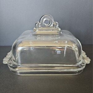 May include: A clear glass butter dish with a rectangular base and a matching lid. The lid features a central arched handle with a circular opening at the top. The edges of the base have a scalloped design, and the glass is smooth and transparent.