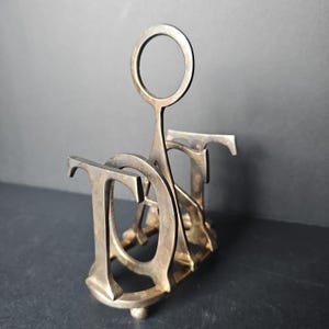 May include: A metallic toast rack featuring a series of interconnected letters forming the word TOAST, with a central vertical handle topped by a circular ring. The structure is crafted from a brass-toned metal with a visible patina and small feet.