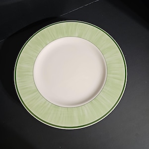 May include: A round ceramic dinner plate featuring a solid cream-colored center and a wide, light green rim. The rim displays a subtle, radiating brushstroke pattern and is finished with a thin, dark green circular border along the edge.