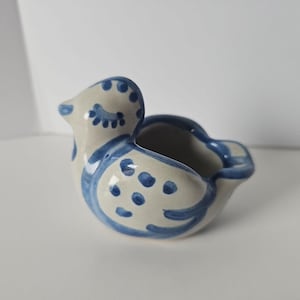 May include: A small, cream-colored ceramic bird-shaped vessel featuring hand-painted blue decorative patterns. The design includes blue dots on the wings, stylized eye markings on the head, and blue outlines along the edges of the bird figure.