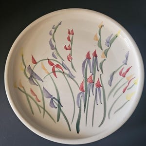 May include: A round, off-white ceramic plate with a hand-painted floral design. The flowers are depicted in shades of red, purple, yellow, and green. The plate has a simple, elegant design, suitable for serving food or as a decorative piece.