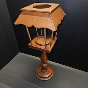 May include: A vintage wooden stand with a square top and a round base. The stand has a decorative, scalloped roof and is supported by vertical rods. The base is round and the stand is made of a medium-toned wood.