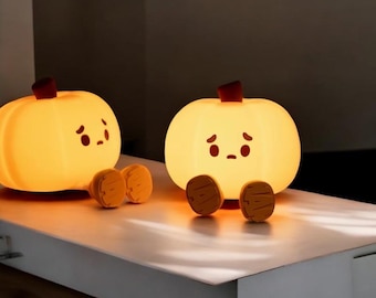 May include: Two glowing pumpkin-shaped lamps with cartoon faces and wooden feet sit on a white surface. The lamps have a warm yellow glow, with a dark red stem on top. The lamps are a cute and whimsical decorative item.