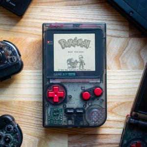 May include: A clear Game Boy Pocket console displaying the Pokémon Red Version game. The console has red buttons and a red directional pad. Other game consoles are visible in the background on a wooden surface.