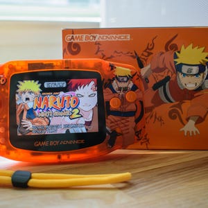 Op de afbeelding: Oranje Game Boy Advance handheld console met een Naruto-game op het scherm. De console heeft een bijpassende oranje polsband. Een doos met Naruto-artwork staat achter de console.