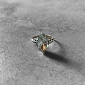 Puede incluir: Un anillo de plata con una gema de corte marquesa y color claro. El anillo tiene un diseño ornamentado en los lados de la gema. El anillo está sobre un fondo gris.