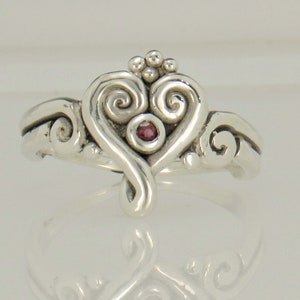 May include: A silver ring with a heart-shaped design featuring a pink gemstone in the center. The heart is adorned with intricate swirls and a crown of small beads.
