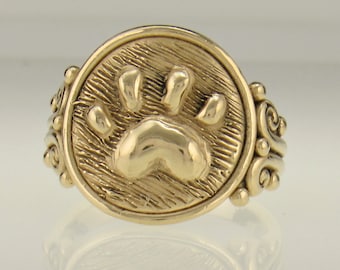 May include: A gold ring with a detailed paw print design on the front. The band is decorated with a swirling pattern.