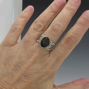 Op de afbeelding: Een zilveren ring met een donkerbruine ovale edelsteen, gezet in een zwierig, versierd ontwerp.