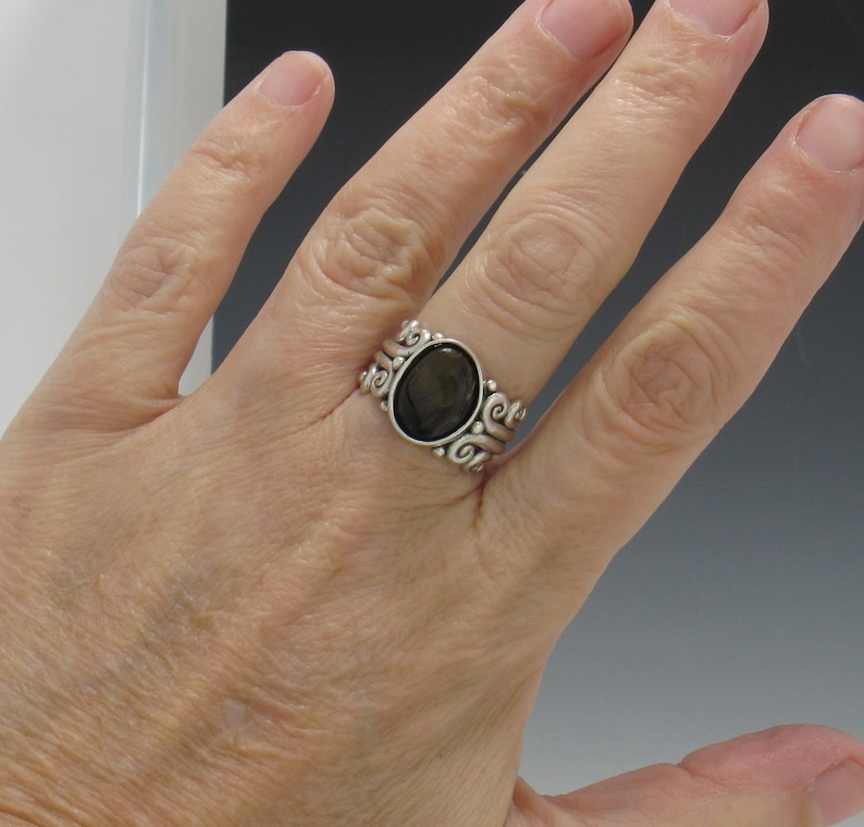 Op de afbeelding: Een zilveren ring met een donkerbruine ovale edelsteen, gezet in een zwierig, versierd ontwerp.