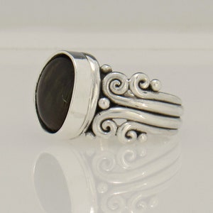 Op de afbeelding: Een zilveren ring met een donkergroene ovale edelsteen gezet in een bezel setting. De band heeft een decoratief ontwerp van draaiende krulletjes.