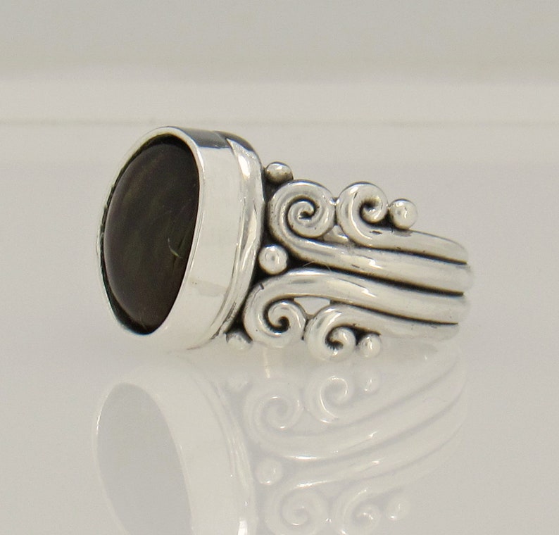 Op de afbeelding: Een zilveren ring met een donkergroene ovale edelsteen gezet in een bezel setting. De band heeft een decoratief ontwerp van draaiende krulletjes.