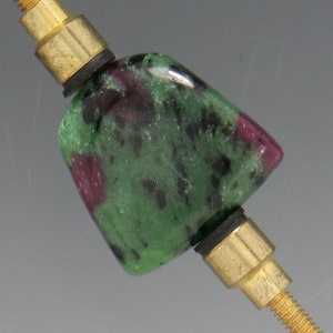May include: A polished gemstone bead with green and red inclusions, mounted on a gold-colored metal rod. The bead is rectangular with rounded edges. The metal rod has a threaded end and a cylindrical section.