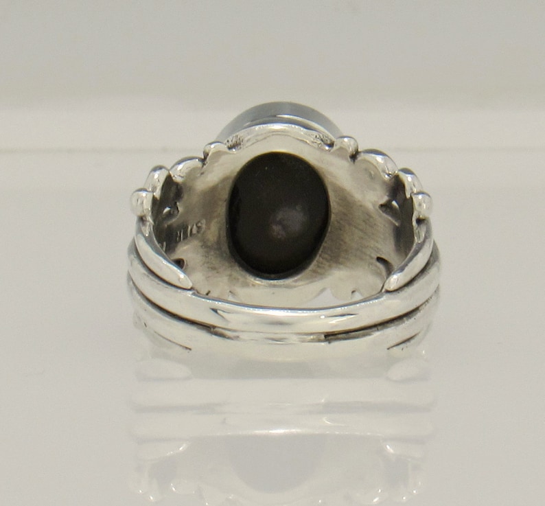 Op de afbeelding: Een zilveren ring met een donkergrijze ovale edelsteen gezet in een gedetailleerd, sierlijk ontwerp. De ring heeft een brede band met een textuur.