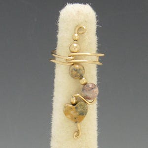 Op de afbeelding: Een goudkleurige draadring met een hartvormige steen en verschillende ronde kralen. De ring is om een vinger-vorm gewikkeld, waarbij het ontwerp wordt getoond. De kralen hebben bruine en paarse tinten. Een sieraad.