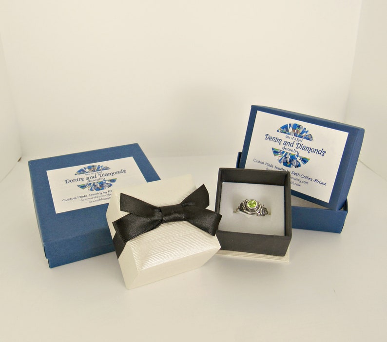 K&ouml;nnte beinhalten: Eine wei&szlig;e Ringbox mit einer schwarzen Schleife steht auf einer wei&szlig;en Oberfl&auml;che neben zwei blauen Boxen. Die Boxen sind mit "Denim and Diamonds Jewelry" beschriftet. Die Ringbox enth&auml;lt einen silbernen Ring mit einem gr&uuml;nen Edelstein.