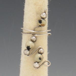 Op de afbeelding: Een zilverkleurige ring met een uniek ontwerp. De ring heeft een reeks kleine, ronde, gespikkelde kralen en zilveren bollen. De kralen zijn licht van kleur met donkere vlekken. De ring is gewikkeld rond een vinger-vormige vorm.