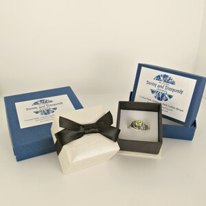May include: A white ring box with a black bow sits on a white surface next to two blue boxes. The boxes are labeled "Denim and Diamonds Jewelry." The ring box contains a silver ring with a green gemstone.
