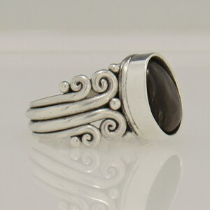 Op de afbeelding: Een zilveren ring met een donkergrijze ovale edelsteen in een bezel-setting. De band heeft een decoratief ontwerp van draaiende lijnen.