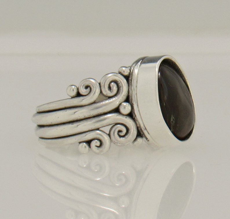 Op de afbeelding: Een zilveren ring met een donkergrijze ovale edelsteen in een bezel-setting. De band heeft een decoratief ontwerp van draaiende lijnen.