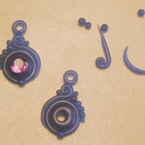 K&ouml;nnte beinhalten: Zwei blaue und violette Wachsohrring-Komponenten mit einem violetten Edelstein in einer der Komponenten. Es gibt auch drei blaue Wachsperlen und zwei blaue Wachswirbel.