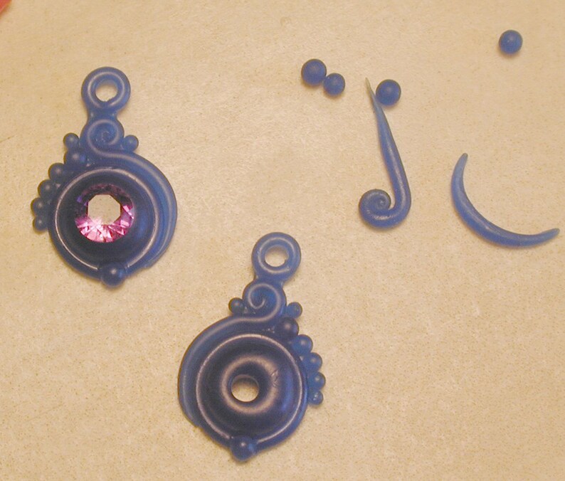K&ouml;nnte beinhalten: Zwei blaue und violette Wachsohrring-Komponenten mit einem violetten Edelstein in einer der Komponenten. Es gibt auch drei blaue Wachsperlen und zwei blaue Wachswirbel.