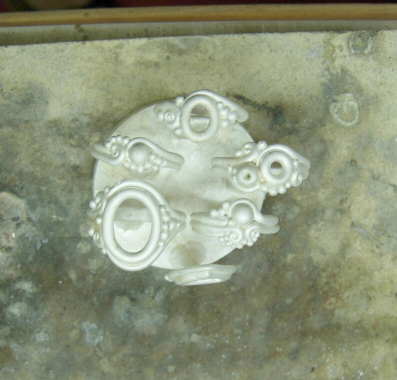 May include: A close-up of six silver rings with different designs. The rings are arranged in a circle on a gray surface.
