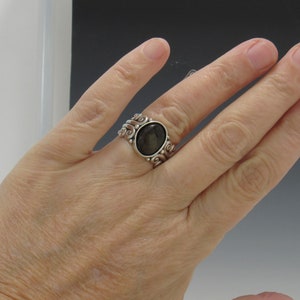 Op de afbeelding: Een zilveren ring met een donkergrijze ovale edelsteen gezet in een wervelend ontwerp.