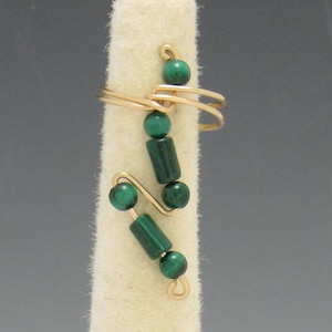 Op de afbeelding: Een goudkleurige draadring met een uniek ontwerp, versierd met donkergroene malachietkralen en cilindrische elementen. De ring wordt gepresenteerd op een witte, cilindrische vorm, die de ingewikkelde, handgemaakte details benadrukt.
