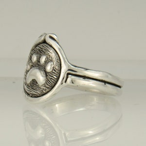 May include: A silver ring with a paw print design. The ring is made of sterling silver and has a textured finish.