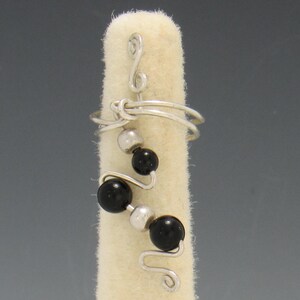 Op de afbeelding: Een zilverkleurige ring met een uniek ontwerp. De ring heeft een reeks zwarte en zilveren kralen, verbonden door een draad die om de vinger gewikkeld is. Het ontwerp omvat een decoratieve krul aan de boven- en onderkant van de ring.