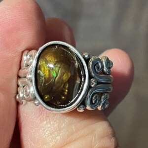 Op de afbeelding: Een zilveren ring met een grote, ovale, iriserende edelsteen die in een bezel-setting is gezet. De ringband is versierd met ingewikkelde krulwerk.