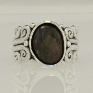 Op de afbeelding: Een zilveren ring met een donkergroene ovale edelsteen, gezet in een zwierig, versierd ontwerp.