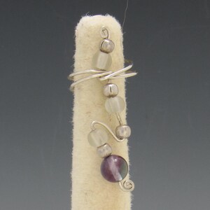 Op de afbeelding: Een zilveren draadring met een decoratief ontwerp. De ring heeft een reeks kleine, heldere kralen en zilveren kralen, samen met een grotere, paarse en iriserende kraal. De ring wordt gepresenteerd op een wit oppervlak.