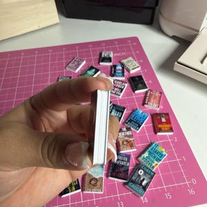 May include: A collection of miniature book replicas with various book titles visible, including "It Starts With Us" and "The Housemaid's Secret." The books are scattered on a pink cutting mat, with a stack of books held in a hand.